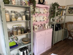 A green shelf unit full of dried goods, a pink dresser with lots of china plates and another green shelf unit