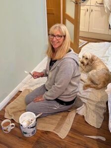 Woman with a paint brush smiling at the camera, with a dog lying behind her. Frugal DIY