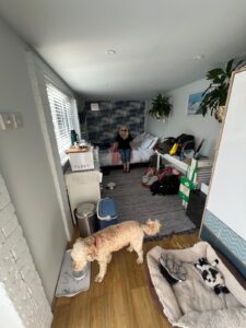 a small room with a woman sitting on a bed and a dog drinking from a bowl