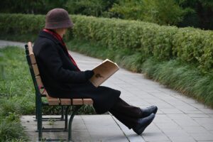 Older person with hat and gloves reading on a bench