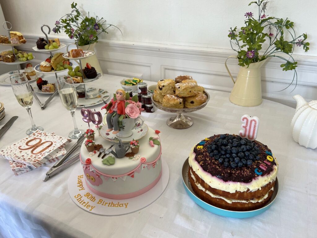 A table with two large decorated birthday cakes, and plates of scones and cakes behind.